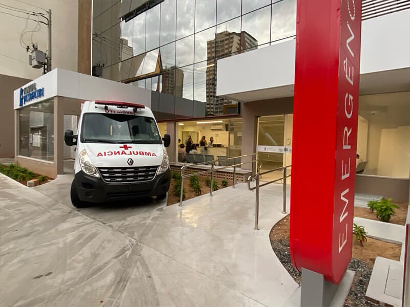Entrada da emergência do hospital, com totem vermelho de sinalização e ambulância estacionada